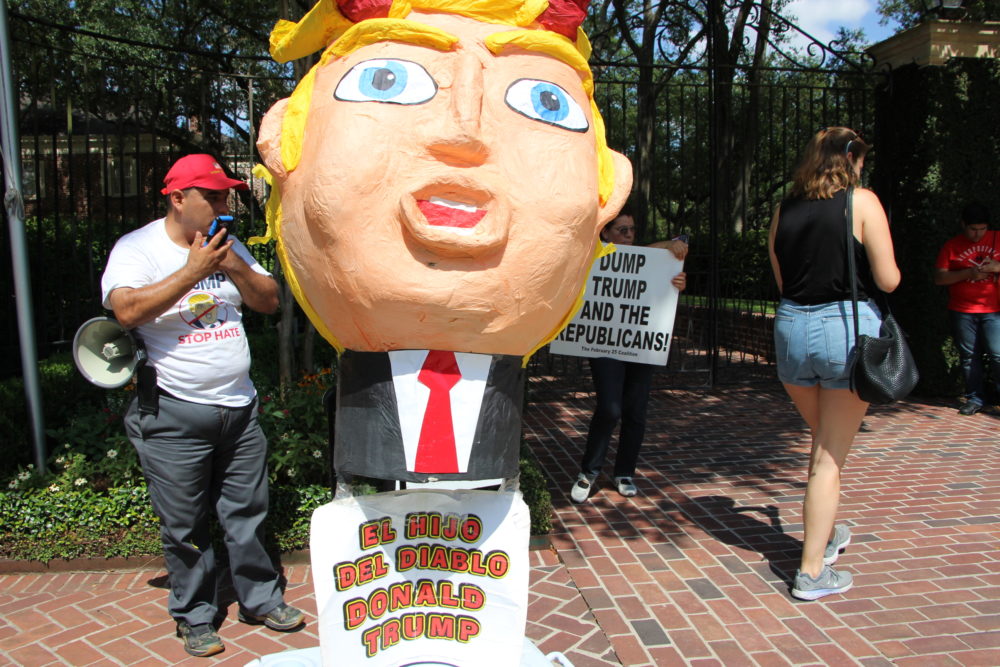 A large paper mache pinata overlooks the sidewalk where protesters are gathered. 